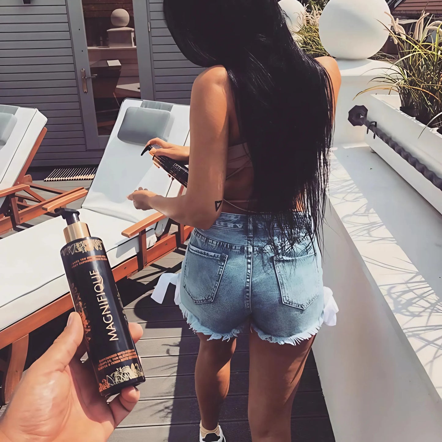Osoba trzyma butelkę balsamu Onyx Magnifique, w tle kobieta w jeansowych szortach i topie przygotowuje leżak na tarasie w słoneczny dzień