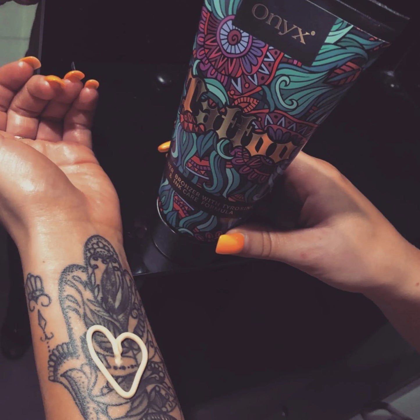 Kobieta z pomarańczowymi paznokciami nakłada balsam brązujący Onyx Tattoo Bronzer na tatuaż na przedramieniu, tworząc kształt serca z kremu pielęgnacyjnego