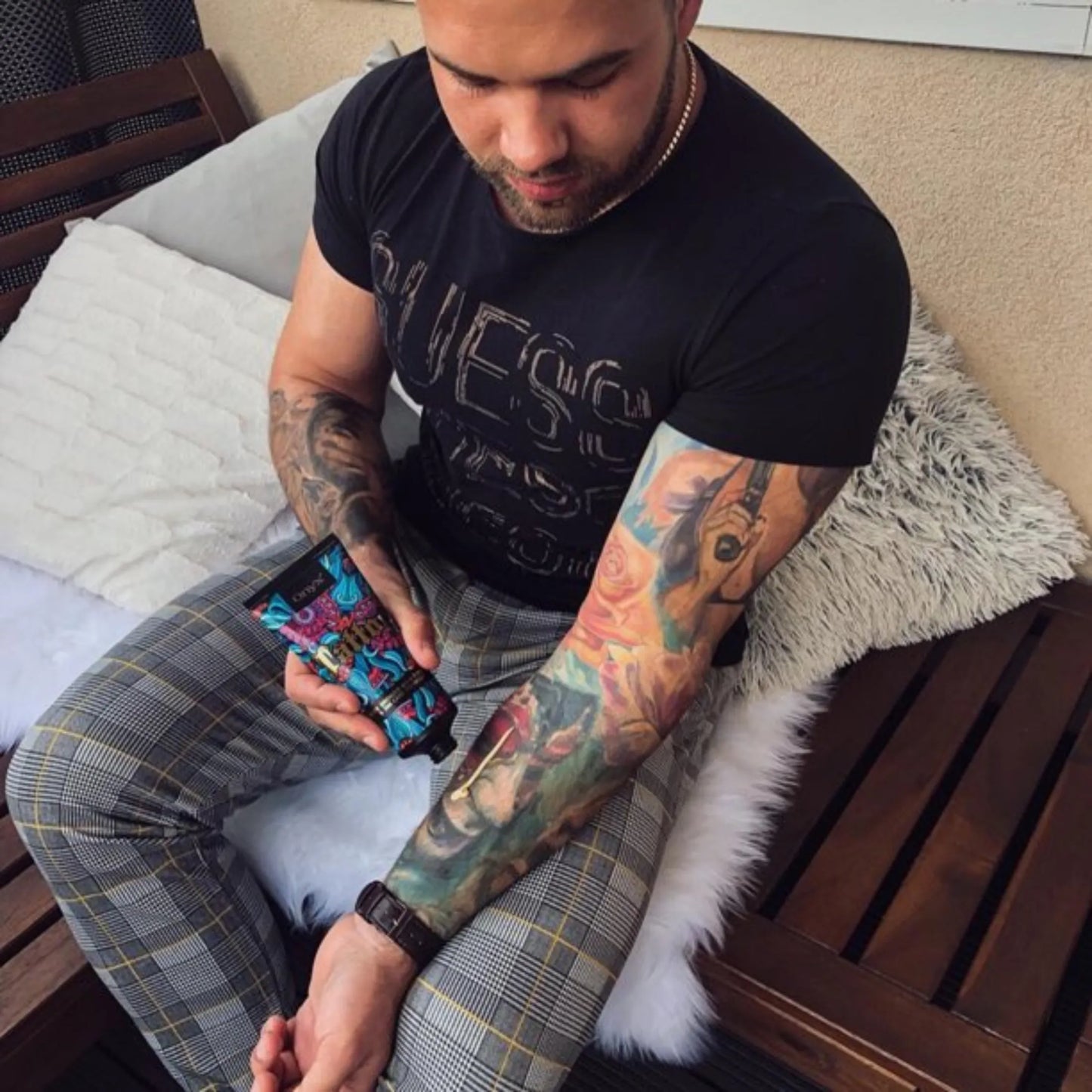 Mężczyzna z kolorowymi tatuażami na ramieniu siedzi na tarasie i nakłada balsam brązujący Onyx Tattoo Bronzer na rękę, podkreślając intensywne kolory tatuażu